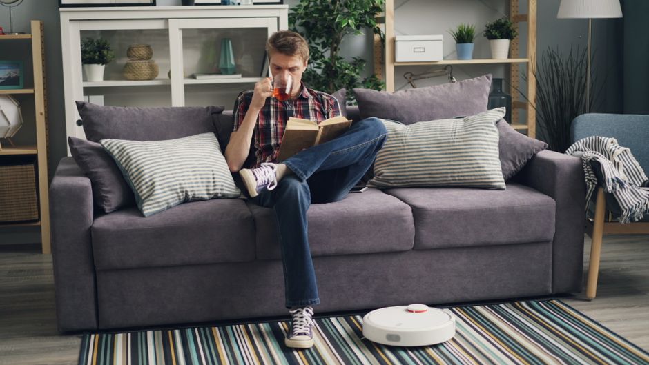Adult man enjoys reading a book and drinking tea in a modern living room, featuring a cleaning robot and sofa.